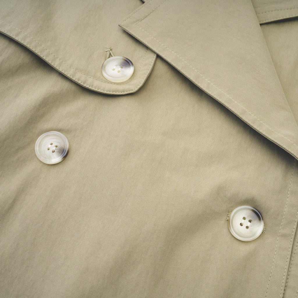 Abrigo trench con cinturón de la marca MRNewyork, de corte clásico y color neutro, con detalles de botones y bolsillos visibles.